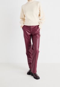 Pull en tricot crème avec un col haut et des poignets côtelés, associé à un pantalon en simili cuir bordeaux et des bottines noires.