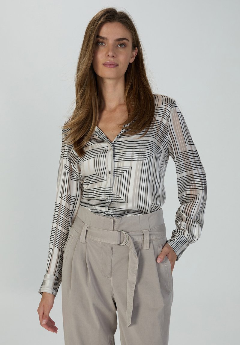 Long-sleeve button-up blouse in white with black geometric print; lightweight fabric with a smooth texture. Paired with high-waisted beige trousers.
