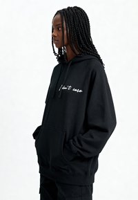 Sweat à capuche noir en tissu doux, avec une poche frontale et le texte "Je m'en fiche" en cursive blanche. Coupe décontractée avec poignets côtelés.