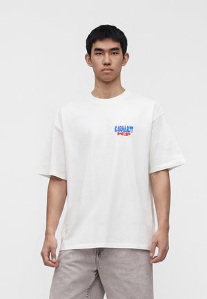 Mladý muž nosí biele oversized tričko s modro-červeným logom Carhartt WIP na hrudi, stojí pred jednofarebným pozadím.