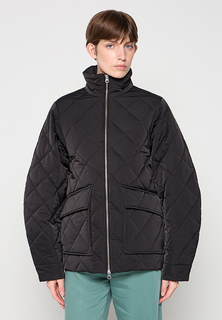 Zwarte gevoerde jas met ritssluiting, hoge kraag en twee voorzakken. Beschikt over een puffy textuur en een moderne uitstraling.