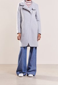 Světlě šedý vlněný kabát s rozparkovým límcem, se zapínáním na zip, bočními kapsami, v kombinaci s modrými širokými džínovými kalhotami a bílými teniskami.