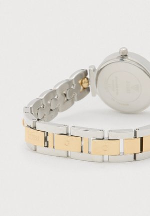 Sudraba un zelta toņa metāla pulkstenis ar saistītu aproces dizainu. Tam ir apaļa korpusa forma ar gludu aizmuguri un gravējumu ar zīmola nosaukumu uz saitēm.