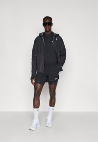 Schwarzes Sportoutfit mit einer Jacke mit Reißverschluss, Shorts und Crew-Socken. Auffällige Nike-Logo-Akzente. Modell trägt sportliche Sonnenbrille und Sneaker.