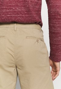 Beige byxor med texturerad yta, med två bakfickor och knappstängning. Bärs med en vinröd långärmad skjorta.