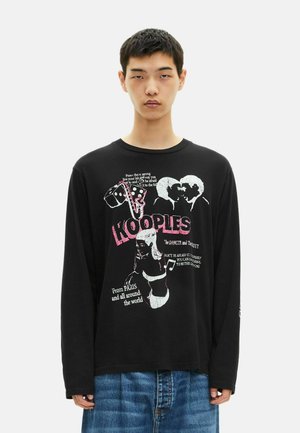 Junger Mann, der ein schwarzes Langarmshirt mit grafischem Text und Bildern trägt, darunter Würfel, ein sich küssendes Paar und eine Frau in Unterwäsche.