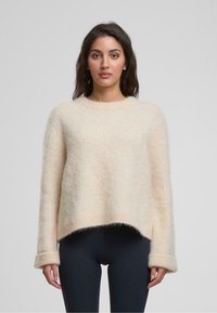 Hellbeige, flauschiger Pullover mit Rundhalsausschnitt, überschnittenen Schultern und gerippten Bündchen. Der Saum ist hinten etwas länger.