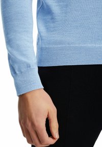 Hellblauer Strickpullover mit taillierter Passform, gerippten Bündchen und glatter Textur, kombiniert mit schwarzen Leggings. Fokus auf Arm- und Ärmeldetails.
