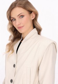 Lys beige læderjakke med høj krave, to foran knapper og en sidelomme, der har en glat overflade og et minimalistisk design.