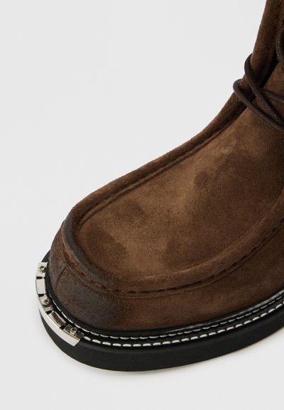 Barna szarvasbőr csizma, lekerekített orral, fekete gumitalppal és ezüstszínű fém részlettel az élén, varrásos díszítéssel.