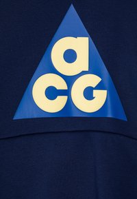 Vestuário azul marinho com um grande triângulo azul e as letras "acg" em negrito, na cor creme. O material parece texturizado e suave.