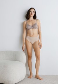 Beige mesh bra with adjustable straps paired with matching high-waisted briefs. Smooth fabric and minimal design, standing against a light wall.