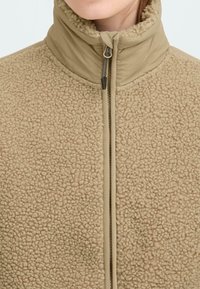 Beige fleecejakke med høj krave og fuld lynlås. Stoffet har en tekstureret, plysset overflade med glatte detaljer på kraven og lynlåsen.