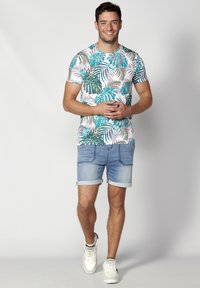 Camiseta con un estampado de hojas tropicales multicolor sobre un fondo blanco, combinada con pantalones cortos de denim enrollados y zapatillas blancas.