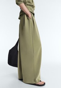Personne portant un pantalon ample vert olive et une veste assortie, tenant un sac noir, debout pieds nus sur un fond blanc uni.