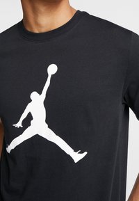 Camiseta de algodón negra con una gran silueta blanca de un jugador de baloncesto saltando con un balón, cuello redondo y mangas cortas.