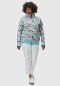 Felpa con zip a fantasia floreale in azzurro chiaro, con colletto alto, polsini e orlo a costine. Indossata con pantaloni bianchi a vita alta e sneakers.