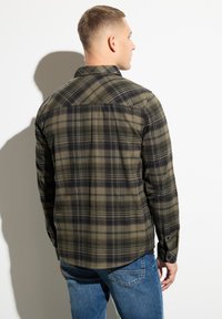 Camisa de franela de cuadros verdes y negros con mangas largas, ajustada en los hombros, dobladillo curvado y puños con botones, combinada con jeans azules.