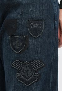 Tamne traperice s tri izvezene zakrpe: štit s natpisom "Diesel", ljiljan i štit s krilima i tekstom.