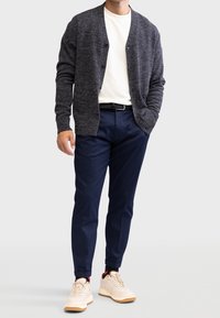 Dunkelgraue Strickjacke, weißes T-Shirt, marineblaue Hose und beige Turnschuhe. Die Strickjacke hat eine strukturierte Strickoptik, einen runden Ausschnitt und eine Knopfleiste vorne.