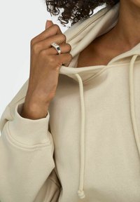 Beige Kapuzenpullover mit hohem Kragen und Kordelzügen. Ribbelbündchen und Saum aus einem weichen, strukturierten Stoff. Hand, die den Kragen greift. Silberner Ring sichtbar.