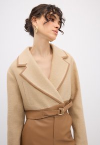 Beige uldblandingsjakke med kontrasterende brune klapper og bælte, med en bred krave og tekstureret stof, parret med en tætsiddende brun nederdel.