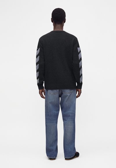 Homme debout, tourné de dos, portant un pull noir avec des bandes en chevron grises sur les manches et un jean bleu ample avec des chaussures noires.