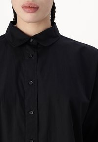 Hedvábná košile na knoflíky z černého materiálu s jemnou texturou, s kulatým límcem a šesti knoflíky vpředu. Jednoduchý design bez viditelných vzorů.