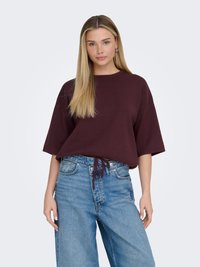 Femme aux longs cheveux blonds portant un haut bordeaux à manches courtes et un jean bleu taille haute devant un fond uni.