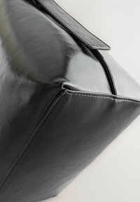 Black leather bag with a glossy finish, rectangular shape, angular corners, and a flat strap. Features visible stitching along the edges.
