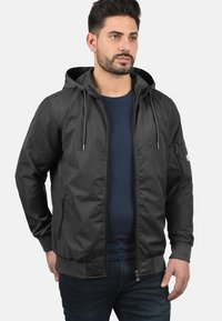 Veste à capuche noire pour hommes en tissu lisse et léger ; présente une fermeture éclair sur le devant, des poignets à bords côtes et une poche sur la manche gauche.