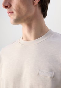 Felpa beige chiaro con scollatura rotonda, texture liscia e un leggero motivo melangiato. Presenta un piccolo etichetta di marca sul petto.