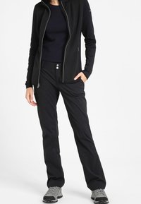 Giacca nera con zip e un motivo testurizzato, abbinata a pantaloni neri slim-fit con due bottoni. Scarpe da escursionismo grigie completano l'outfit.