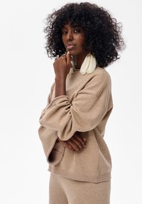 Beżowy sweter o luźnym kroju, wykonany z teksturowanego, błyszczącego materiału, z szerokimi rękawami. Modelka ma kręcone włosy i nosi wyraziste kolczyki.