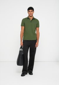 Grønn polo skjorte med teksturert overflate, korte ermer og kontrastkrage, kombinert med svarte bukser og en svart tote bag.