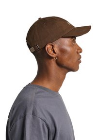 Brown adjustable cap made of fabric with a curved visor. Features a buckle strap, side ventilation holes, and logo embroidery on the front.