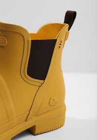 Bota de goma amarilla con un lado elástico de color marrón oscuro, superficie texturizada y un logo prominente en el lateral. Tacón reforzado para mayor durabilidad.