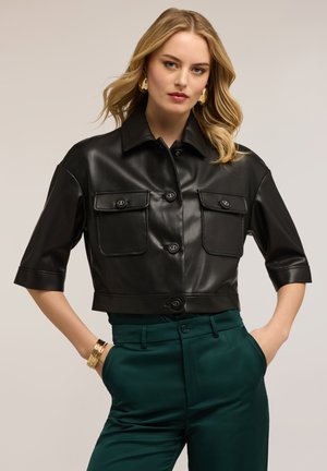 Blonde Frau trägt ein schwarzes, kurzes Lederhemd mit geknöpften Klappentaschen und dunkelgrünen, hoch taillierten Hosen, Hände in den Taschen.