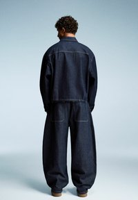 Giubbotto in denim e pantaloni oversize di colore blu scuro. Il giubbotto presenta un colletto classico e due tasche posteriori; i pantaloni hanno una vestibilità rilassata con cuciture visibili.
