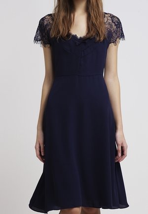 Femme portant une robe bleu marine longueur genoux avec des manches courtes en dentelle et un décolleté en V subtil.