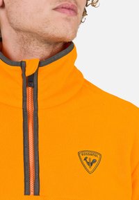 Oranger Fleece-Pullover mit halbem Reißverschluss, grauen Akzenten und einem Rossignol-Logo auf der Brust. Weicher Stoff und hoher Kragen.