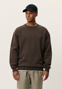 Καφέ φούτερ με ριμπ μανσέτες και τελείωμα, με στρογγυλή λαιμόκοψη και διακριτική κεντητή λογότυπο. Συνδυάζεται με μαύρο καπέλο και ανοιχτόχρωμα σορτς.