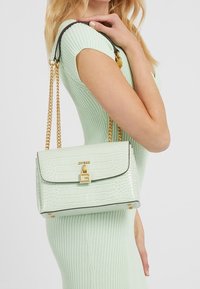 Mátově zelená kabelka z umělé kůže s krokodýlovým embosováním, se zlatými detaily, řetízkovým popruhem a černými prvky. Má klopový uzávěr a logo detail.