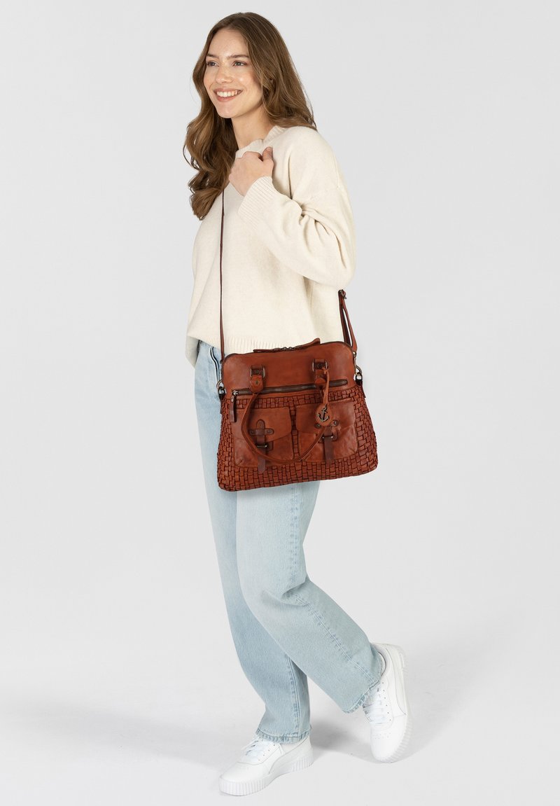 Borsa a spalla in pelle marrone con texture intrecciata, multiple tasche esterne e tracolla regolabile. Abbinata a jeans blu chiaro e sneakers bianche.