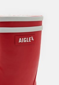 Botte en caoutchouc rouge avec une bande blanche près du haut et une étiquette rectangulaire blanche avec "AIGLE" embossé en lettres rouges.