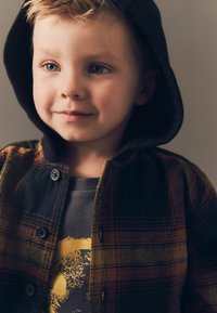 Fliisilest särgiga, millel on must kapuuts, esindades soojaid värve koos ruudulise mustriga. Allpool hall t-särk, millel on ümmargune graafiline kujundus.