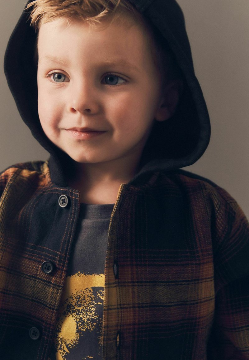 Fliisilest särgiga, millel on must kapuuts, esindades soojaid värve koos ruudulise mustriga. Allpool hall t-särk, millel on ümmargune graafiline kujundus.