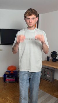 Vaaleanharmaa lyhythihainen t-paita, siniset farkut, pitää laseja kädessään, seisoo sisätiloissa, taustalla näkyy huonekaluja ja kaiutin.