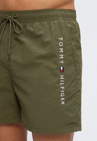 Κοντινό πλάνο ελιάς-πράσινων μαγιό σορτς με κορδόνι στη μέση και κάθετο λογότυπο Tommy Hilfiger μαζί με τη σημαία στη δεξιά πλευρά.