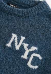 Blauwe gebreide trui met een zachte textuur, een ronde halslijn en de letters "NYC" in contrasterend wit garen. Merkenlabel zichtbaar bij de hals.